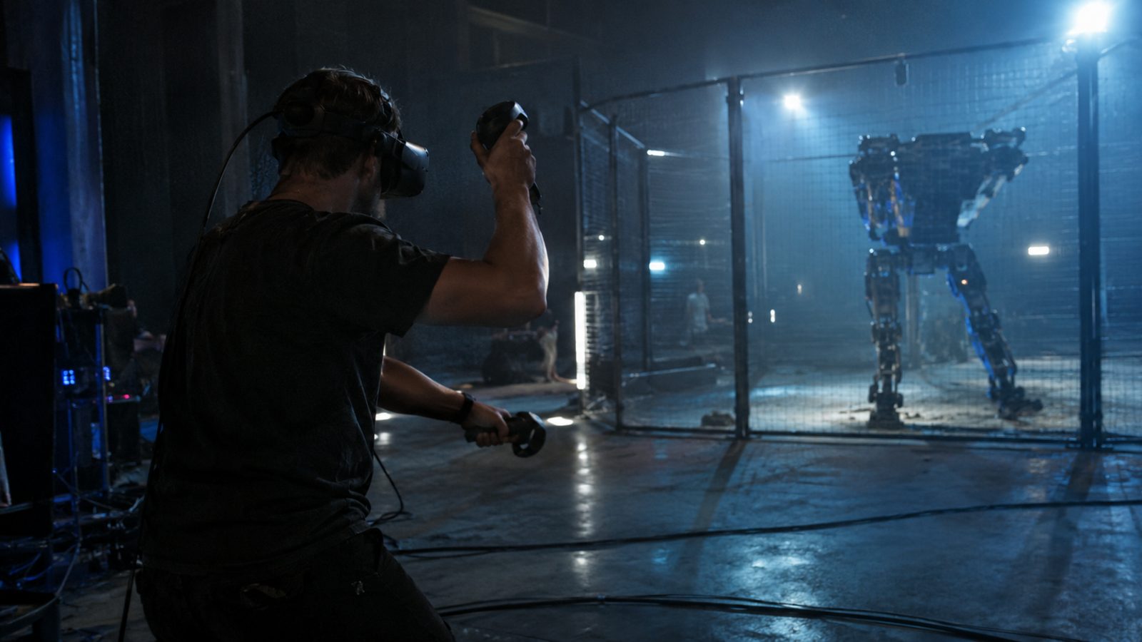 A man in a VR headset holding controllers throws a punch offstage while a lit cage glows behind him