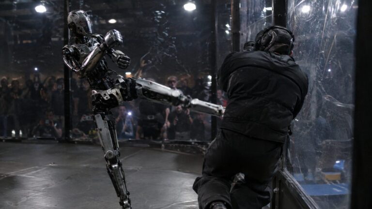 A six-foot metal humanoid robot extends a front kick into a padded man against the clear wall of a fighting cage