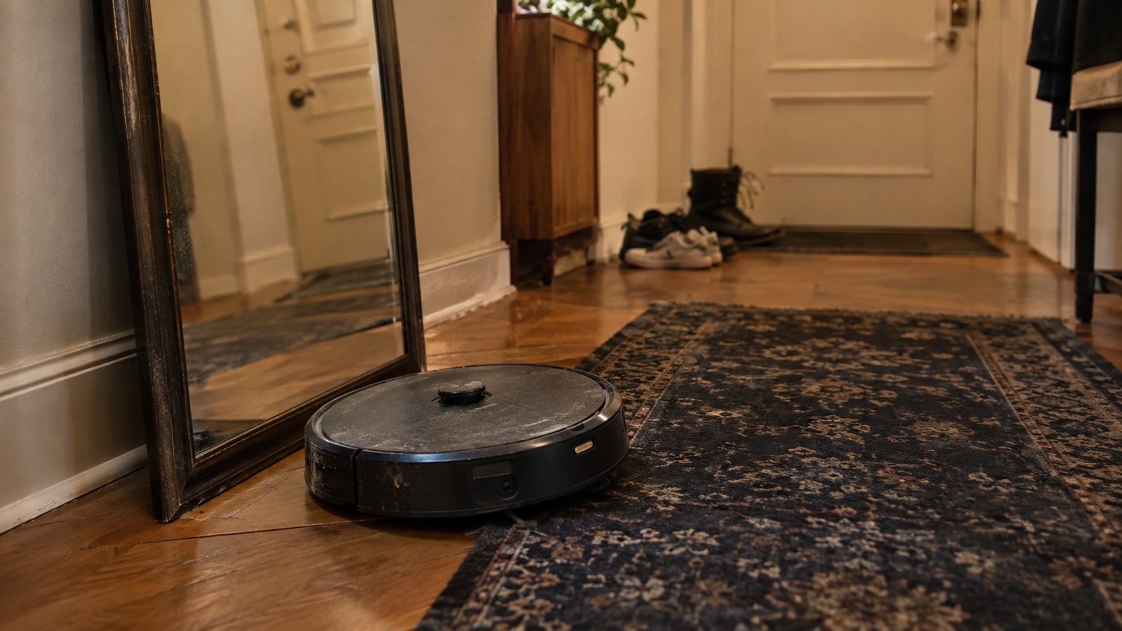 A robot vacuum stopped at the edge of a dark rug in front of a floor-length mirror