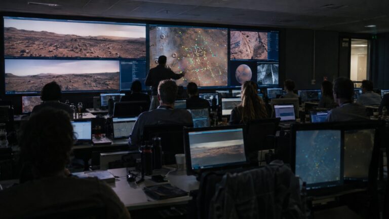 Mission planners seated at rows of desks facing wall screens showing a Mars landscape and target markers