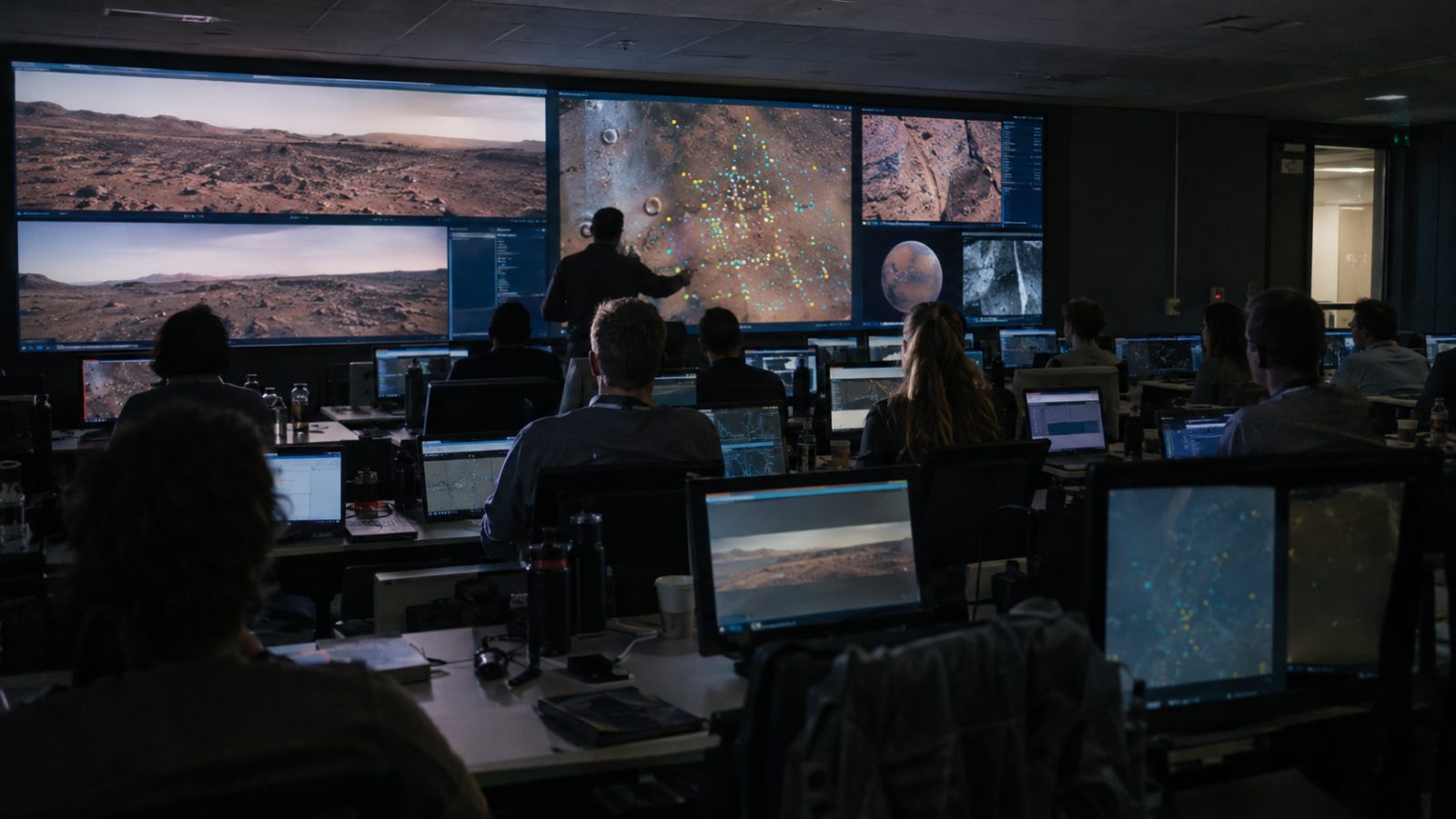 Mission planners seated at rows of desks facing wall screens showing a Mars landscape and target markers