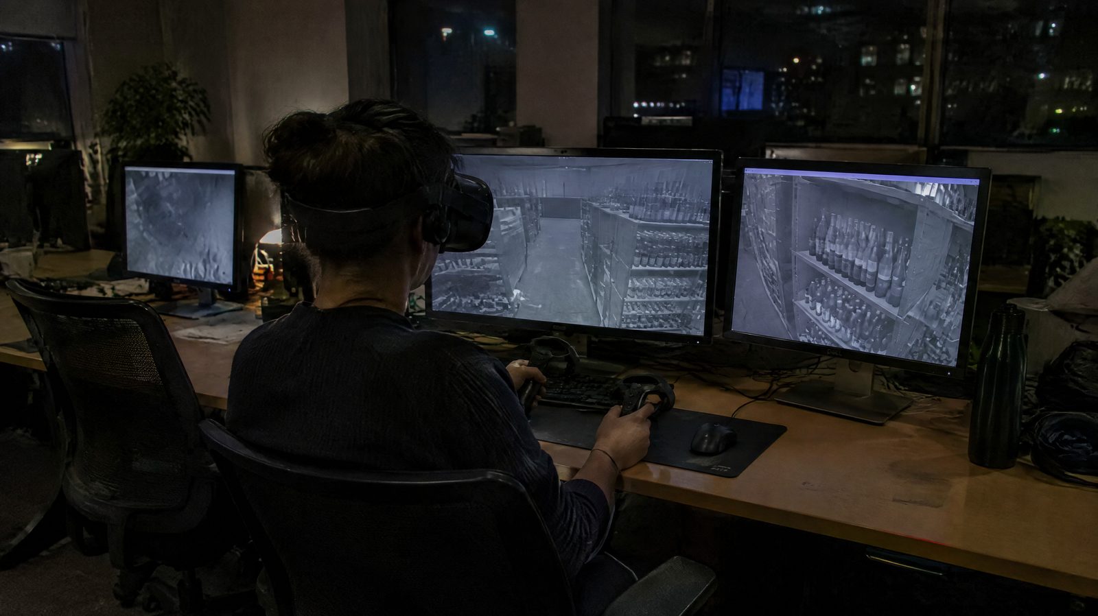 A remote operator seen from behind wearing a VR headset in front of monitors showing a stockroom