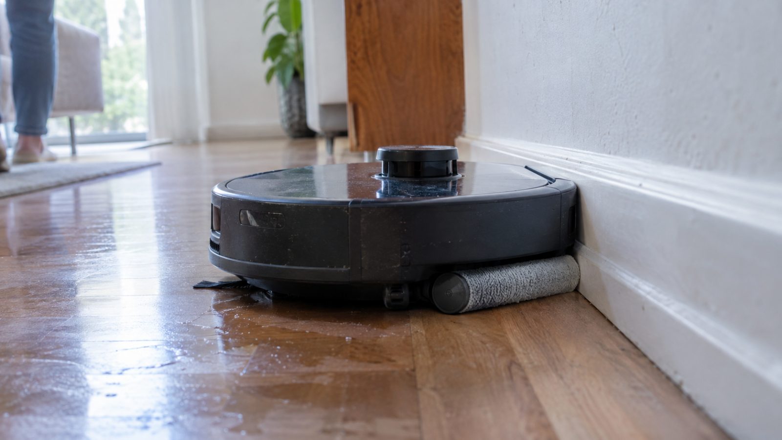 Robot vacuum with a sideways-sliding roller mop cleaning along a baseboard on wood flooring