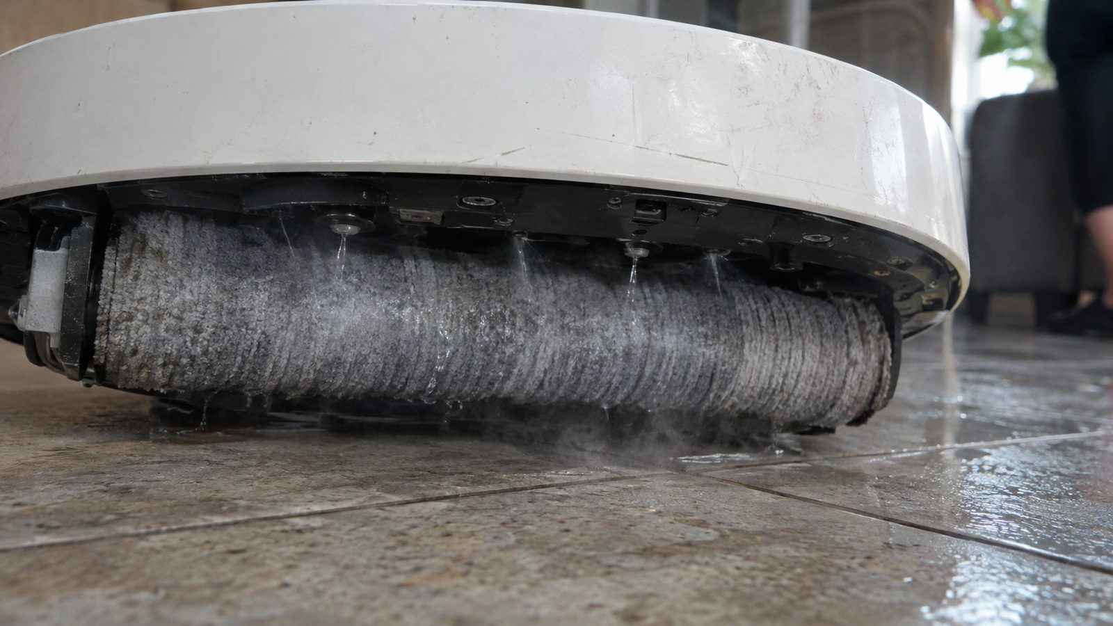 Close-up of a wide flat cloth roller under a robot mop being sprayed with fine jets of hot water