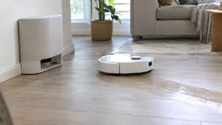 Round white robot vacuum and mop cleaning a wood-look floor, leaving a damp stripe, with its tall white dock against the wall