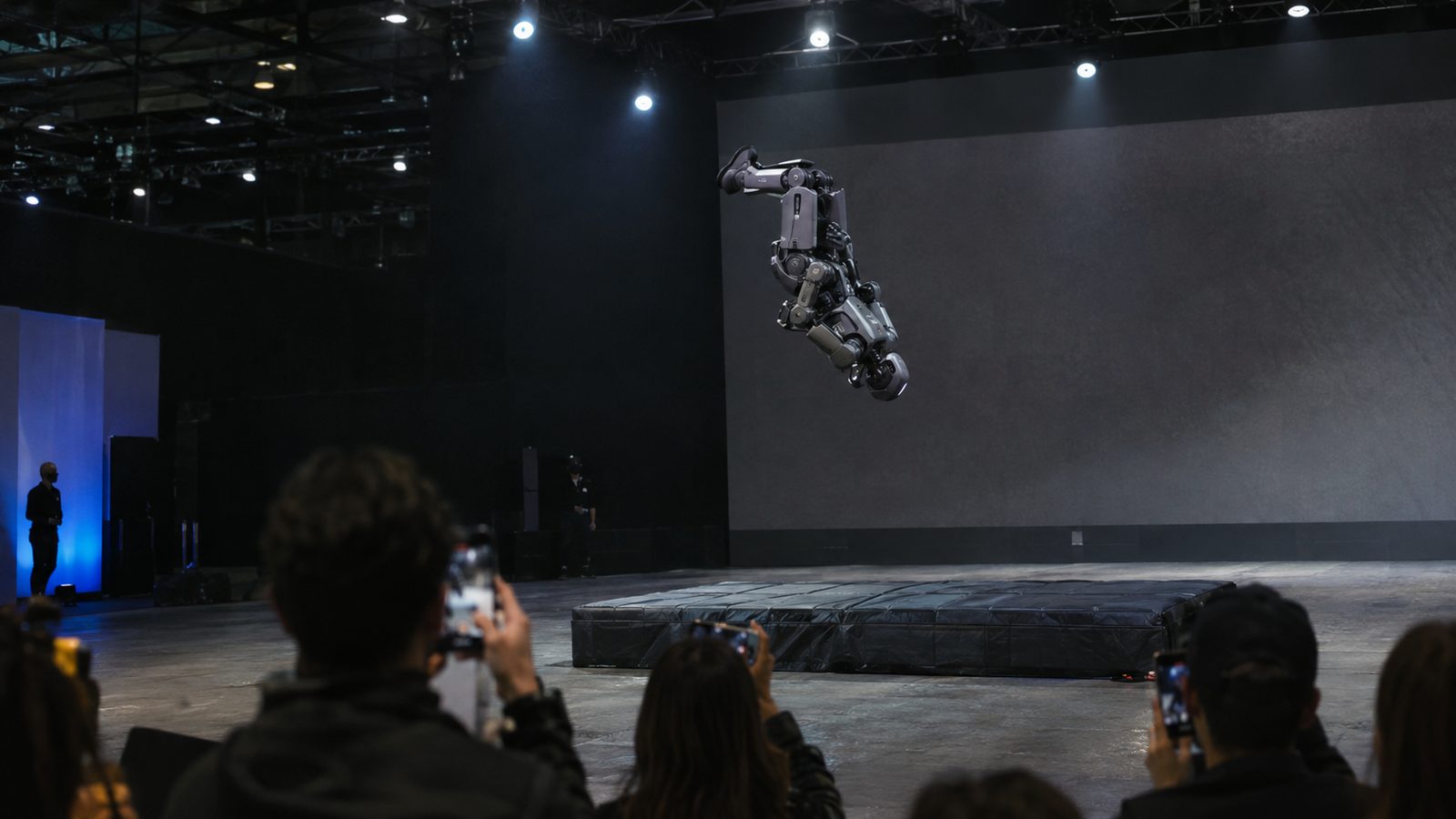 A humanoid robot in the middle of a sideways flip above a black mat, filmed by an audience