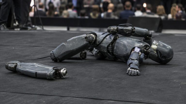 A tall humanoid robot lying on its side on a black mat, its detached lower leg beside it