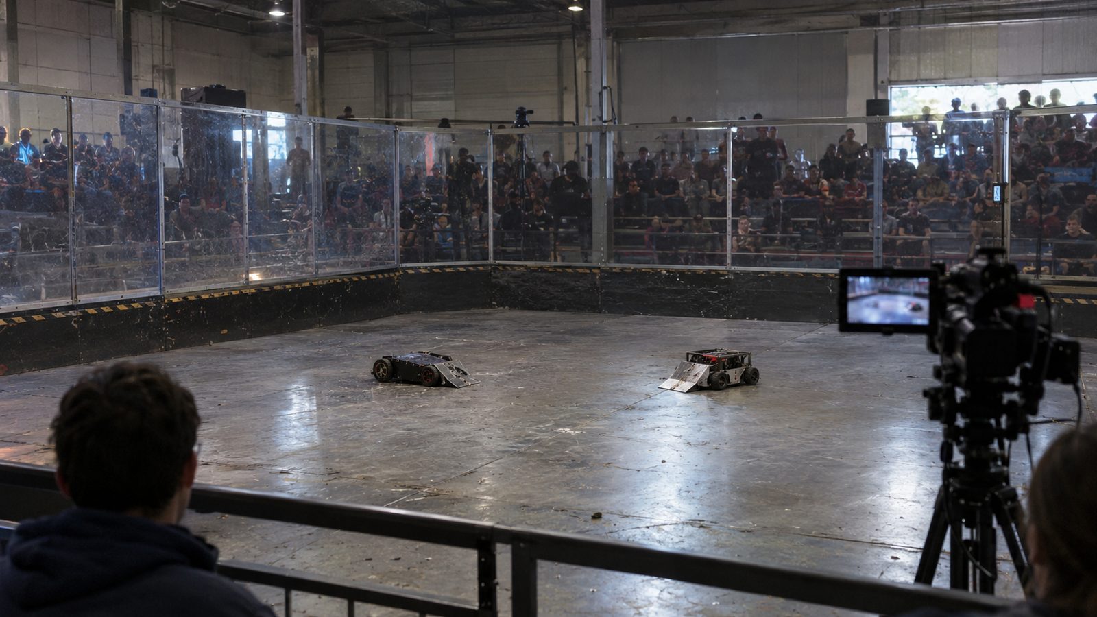 Spectators sit outside an enclosed robot-combat arena while two small robots face off inside.