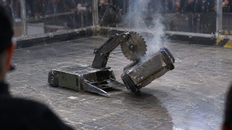 A small combat robot lifts a rival on front prongs and brings an arm-mounted saw down into its lid, releasing smoke inside an enclosed arena.